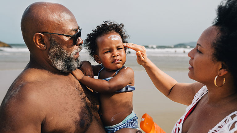 Little girl being held in her father's arms with her mother putting sun block on her face during a family beach vacation in summer; Credit: Giselleflissak