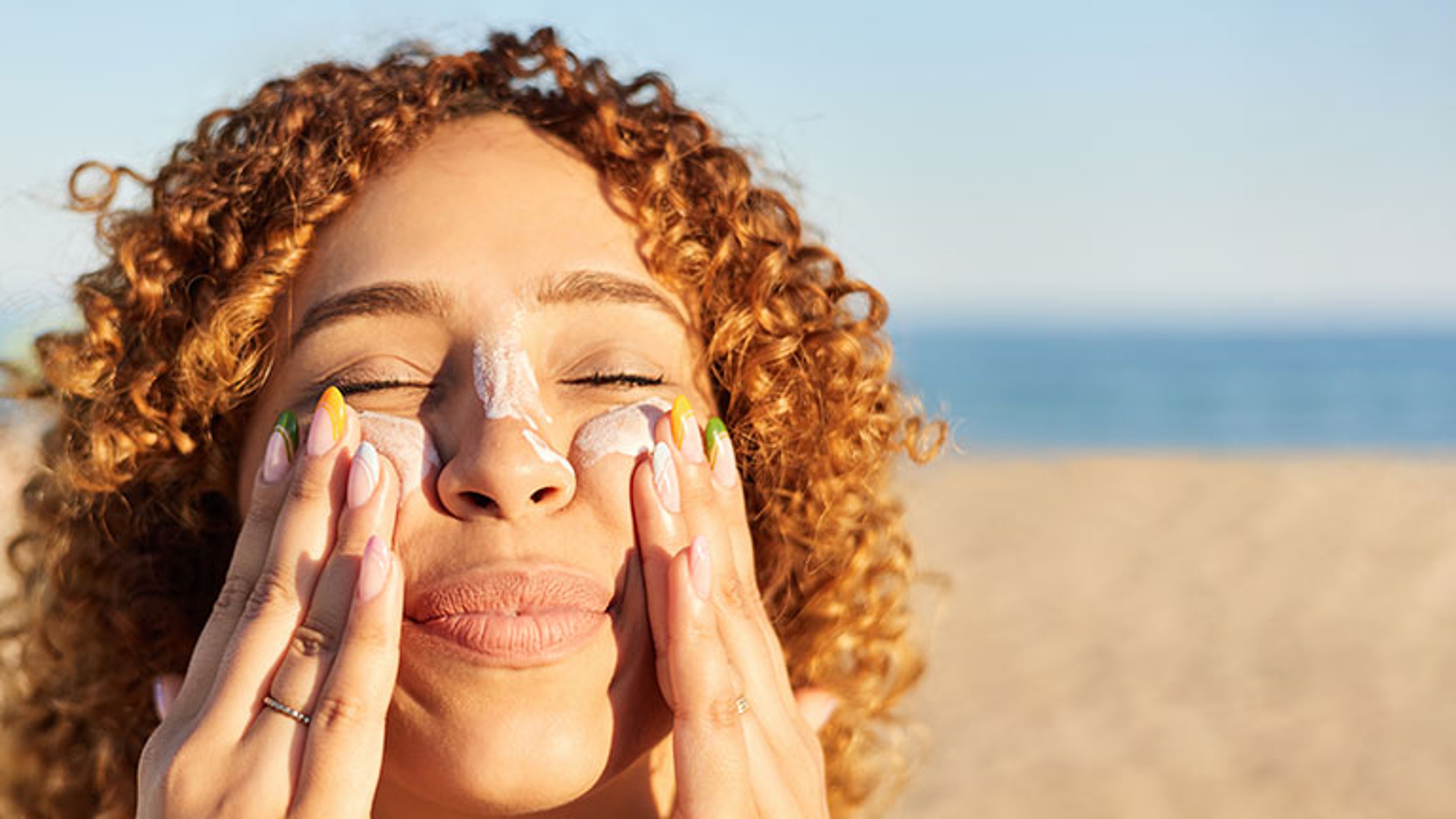 Young latin woman applying sunscreen to her face on the beach in a summer sunset. Cheerful gesture with her eyes closed; Credit: Javi Sanz