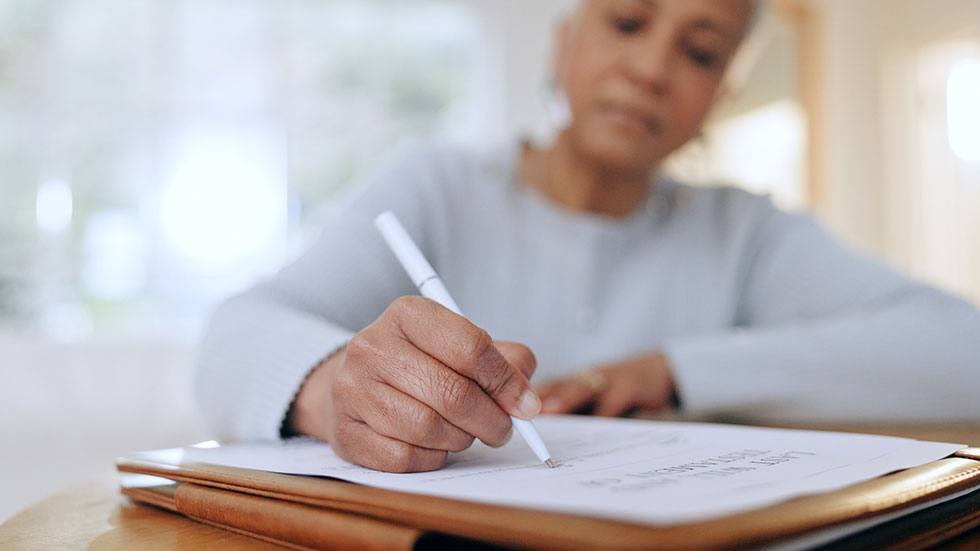 Senior woman, sign and writing with documents, paperwork and application for life insurance policy. Person, hand and checklist for compliance, investment or will in retirement with signature in home.  Credit: Jacob Wackerhausen