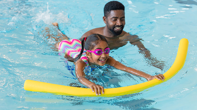 A man teaching his young daughter how to swim in a swimming pool in Boldon, North East England. The daughter is wearing goggles and is using a woggle to help support her in the water.   Credit:SolStock 