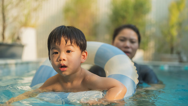 Happy Asian toddler practicing swimming in a blue pool with a safety float ring while playing with a parent. Joyful family bonding moment highlighting early childhood development, muscle strengthening, water confidence, emotional growth, healthy lifestyle