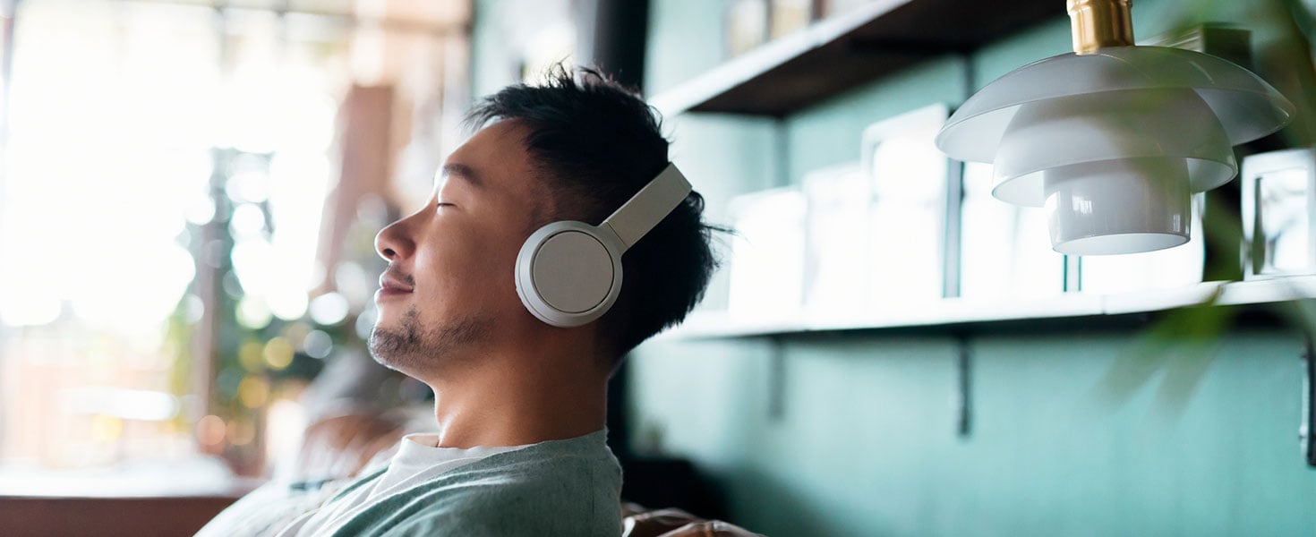 Young Asian man with eyes closed, enjoying music over headphones while relaxing on the sofa at home Credit:AsiaVision