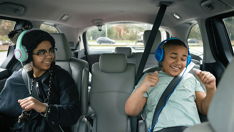 Two multiracial children enjoy a ride to school in the family car, a spacious minivan that accommodates their transportation needs. They listen to music through their headphones, pretend dancing in the car. Credit:RyanJLane