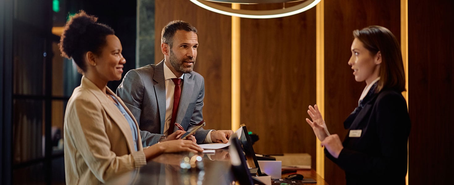 Happy businessman and his female colleague communicating with hotel manager at reception desk; redit: Drazen Zigic