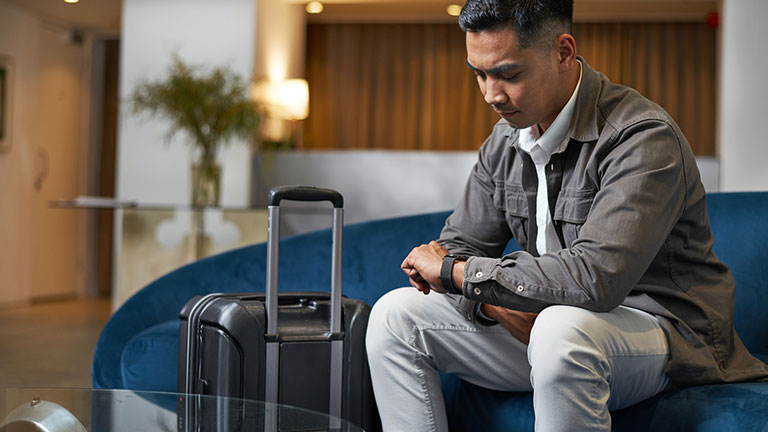 A traveler in a stylish hotel lobby sits on a sofa with luggage nearby, checking the time. The ambiance exudes modernity and professionalism, suggesting a business trip or vacation. Credit: Meeko Media