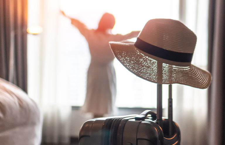hat on top of suitcase handle in hotel room, woman in background stretching near window