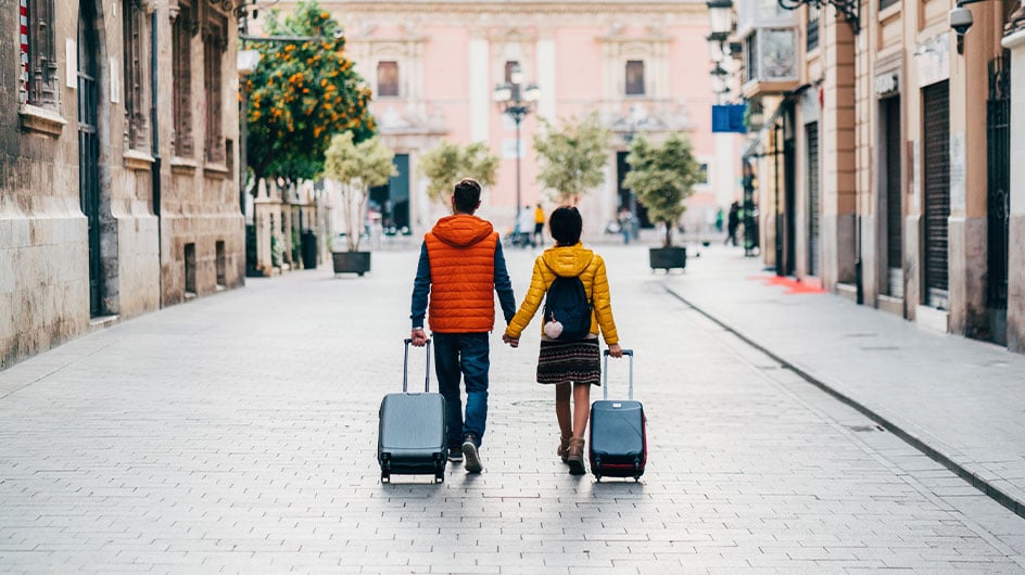 back of man and woman walking, holding hand, and pulling suitcases