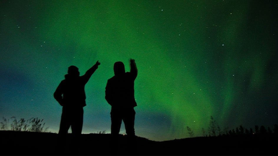silhouette of two people pointing towards northern lights
