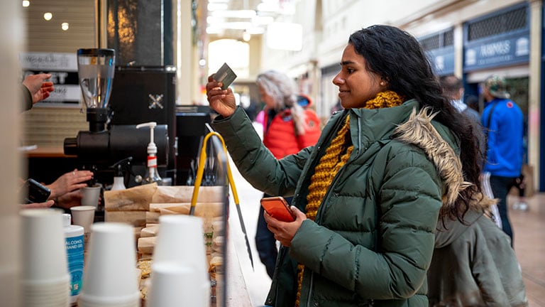 A mid adult woman on a staycation in the city of Newcastle on a sunny winters day. She is wearing casual, winter clothing and accessories. She is paying for a coffee with her credit card at a small coffee shop in a shopping arcade. Credit: SolStock