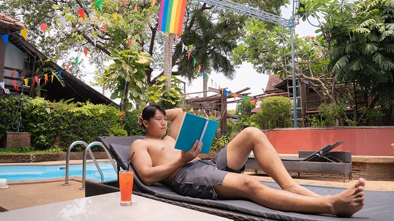 Young man is lying on a sunbed by the pool, engrossed in a book, with a colorful cocktail at his side, while a rainbow flag and festive decorations create a vibrant backdrop See Less By Tj
