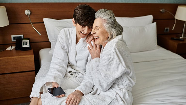 Affectionate senior lesbian couple sitting on top of hotel bed, sharing a loving moment. By Bliss