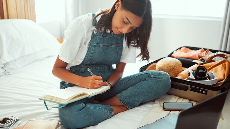 Travel, planning and woman in bedroom with suitcase for summer, vacation or holiday information, checklist and website. Location, hotel and journal book of girl writing notes with clothes in luggage. By Kay A/peopleimages.com
