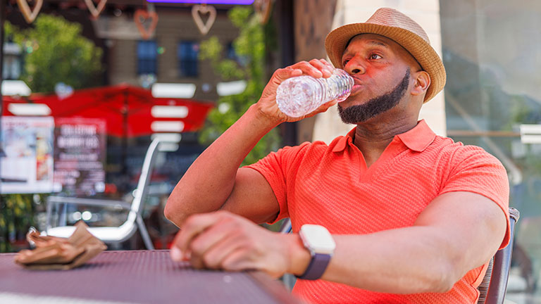 Man in a salmon-colored shirt and straw hat is drinking water from a plastic bottle while sitting at an outdoor cafe table in Madison, NJ. The background shows an open sign and urban surroundings typical for a street cafe. Credit: Alex Potemkin 