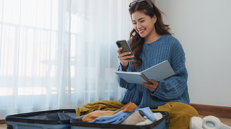 Young asian woman sitting on the floor, using her smartphone while preparing clothes for travel. Checking her list in a notebook, she organizes essentials before packing her suitcase See Less By crizzystudio
