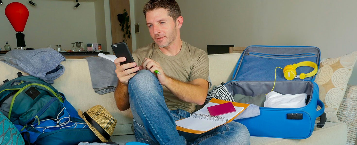 Young handsome and happy man packing travel suitcase at home sofa couch using mobile phone organizing holidays trip booking online smiling cheerful in summer vacation and wanderlust; by Marcos Calvo