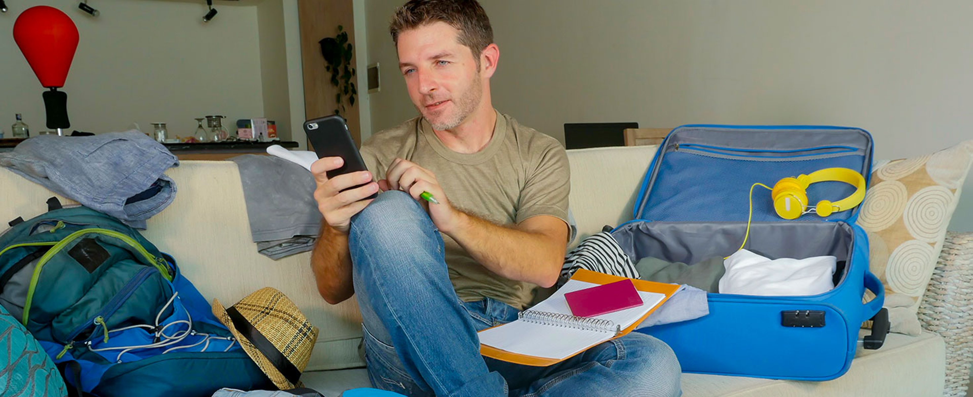 Young handsome and happy man packing travel suitcase at home sofa couch using mobile phone organizing holidays trip booking online smiling cheerful in summer vacation and wanderlust; by Marcos Calvo
