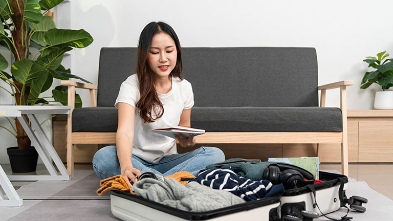 Travel Preparation. A woman sits on the floor, reviewing her packing list while organizing her suitcase. By wichayada