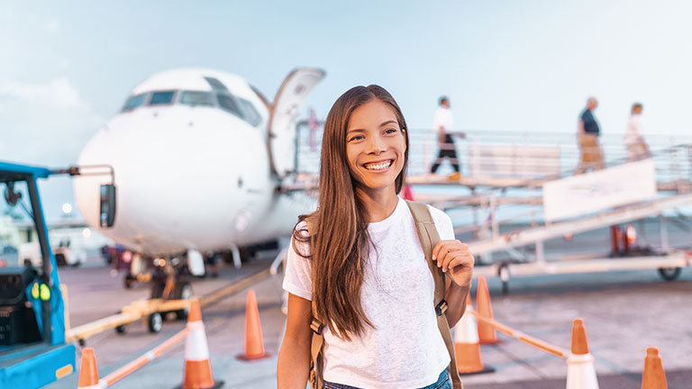 Travel woman arriving at destination coming out of plane after flight at airport. Happy young Asian tourist girl walking on outside tarmac for vacation business trip by Maridav