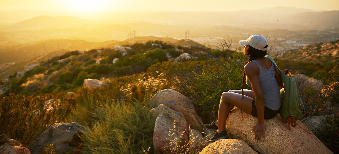 Rear view of woman hiker sitting on rock on top of hill while looking at sunset over San Diego California