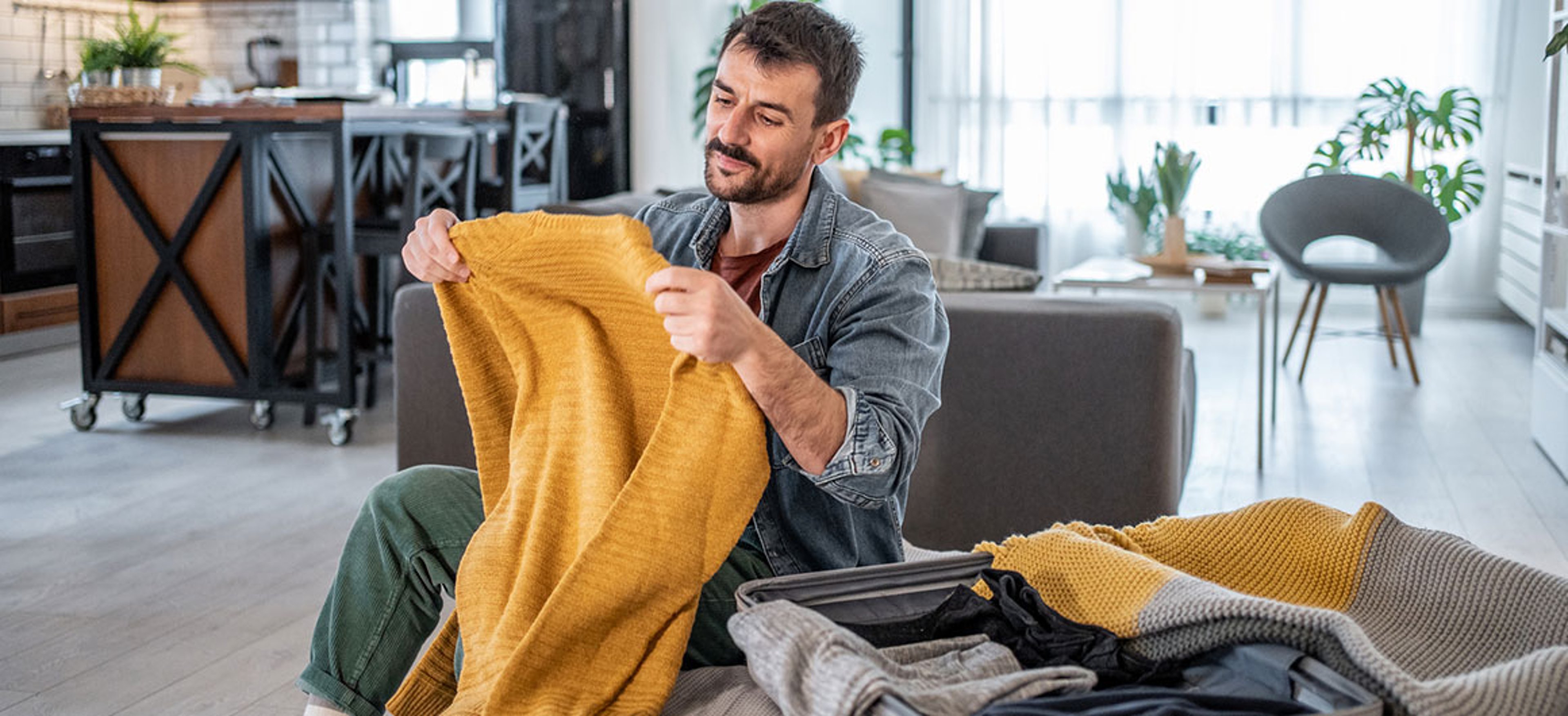 Young man is sitting on a sofa in his modern apartment, carefully folding and packing a yellow sweater into his suitcase, preparing for an upcoming trip