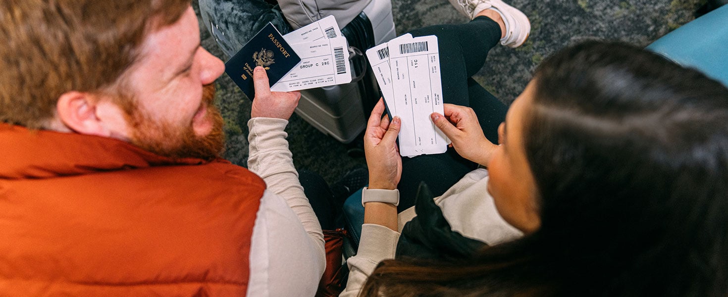 Excited Married Couple Sitting Together in an Airport Departure Area, holding Airplane Flight Tickets & Passports, Ready to Travel Together for Holiday Credit:Jeremy Poland