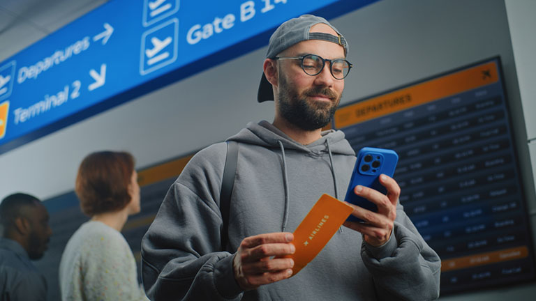 Crowded Airport Terminal: Happy Man Scanning Plane Ticket Using Smartphone to Check Flight Information. Digital Arrival and Departure Information Screen in Background. Traveler Going on Holiday Trip Credit:EvgeniyShkolenko