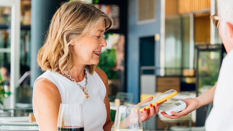 Elegant mature woman paying wirelessly at restaurant terrace, holding wine glass near smiling waiter processing contactless payment with pos termina by Dani D.G