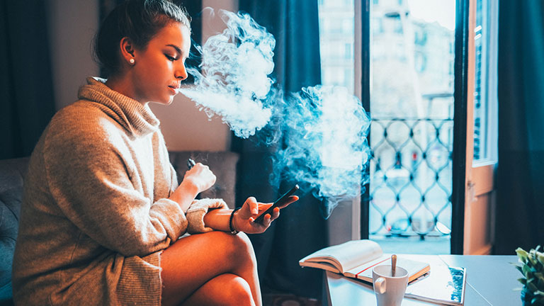 Tourist woman sits comfortably inside her apartment in Barcelona, exhaling vapor from her electronic cigarette while using her smartphone. The cozy atmosphere, open window, and city views embody a relaxed travel moment in Spain. Credit:martin-dm