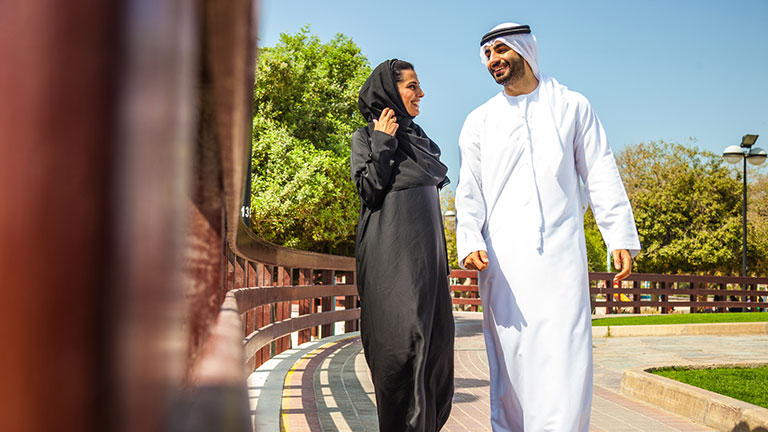 Traditional Emirati young couple enjoying life outdoor during a sunny day. The man in front wearing the dishdash traditional wear, the woman wearing the traditional abaya all black dress. Credit:LeoPatrizi