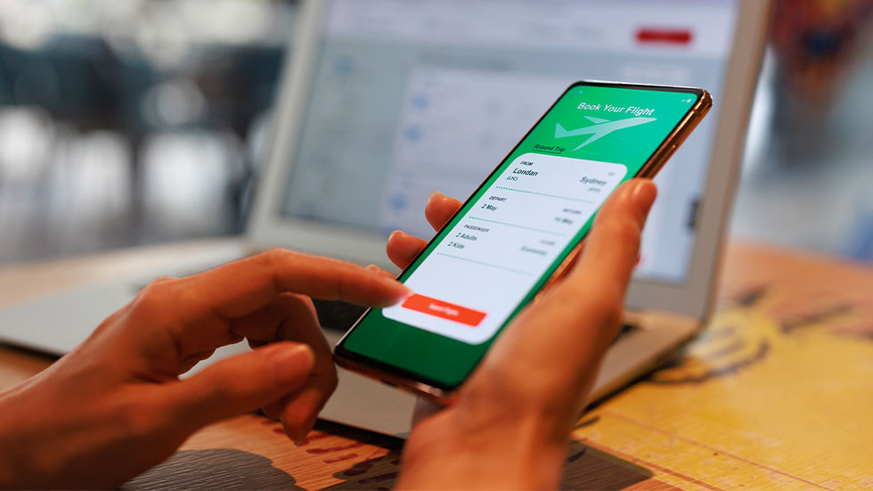 person booking flight on phone, laptop screen in background