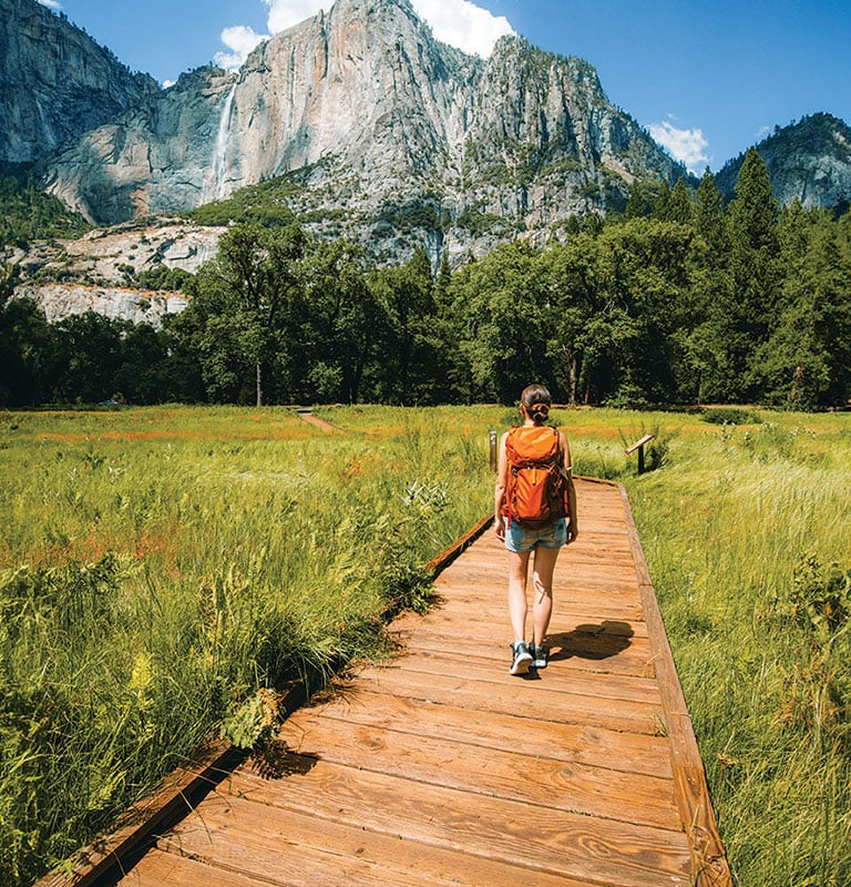 Young woman wearing a backpack is walking on a wooden trail towards the Yosemite falls near Merced river in Yosemite valley, Yosemite National Park in United states of America; photo by Victoria Nefedova
