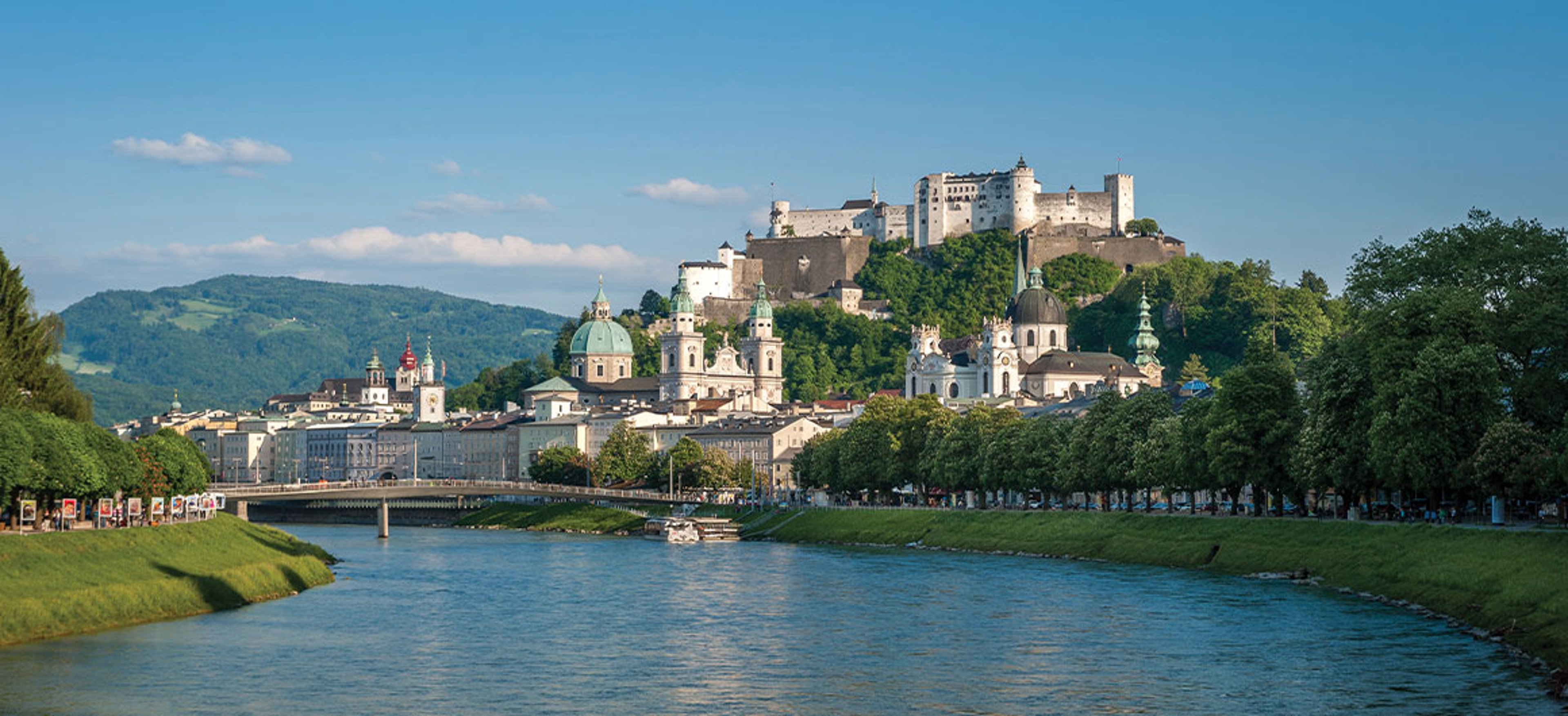 Scenic view of Salzburg, Austria, featuring the historic Hohensalzburg Fortress, baroque architecture, and the Salzach River with lush greenery under a blue sky.