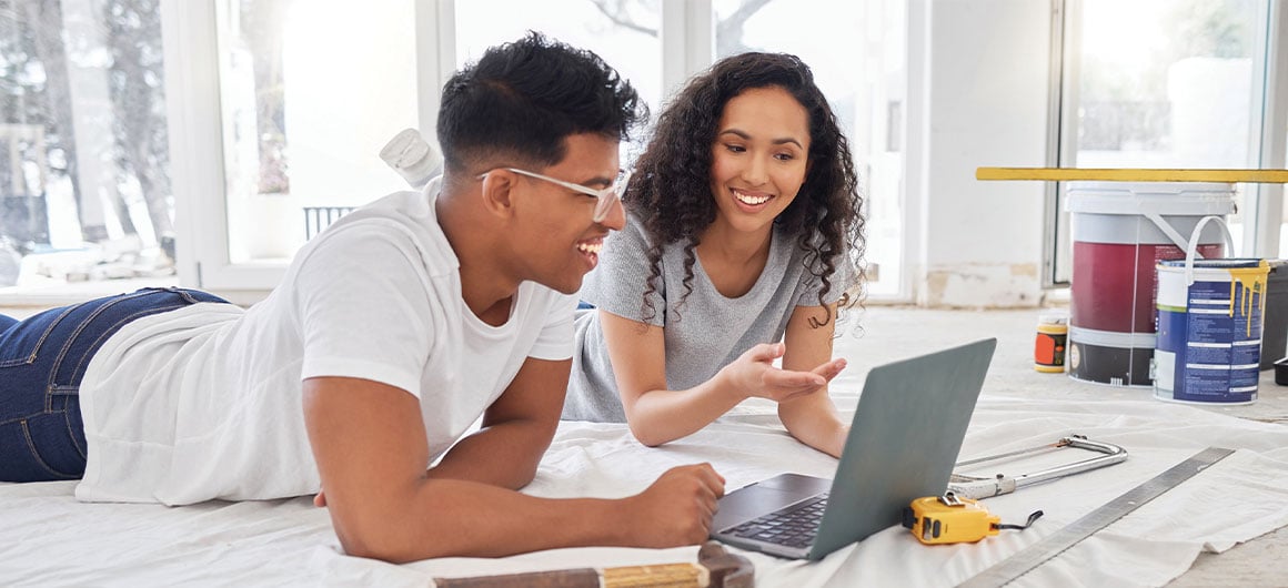 man and woman looking at laptop, laying on floor, in the midst of a home reconstruction