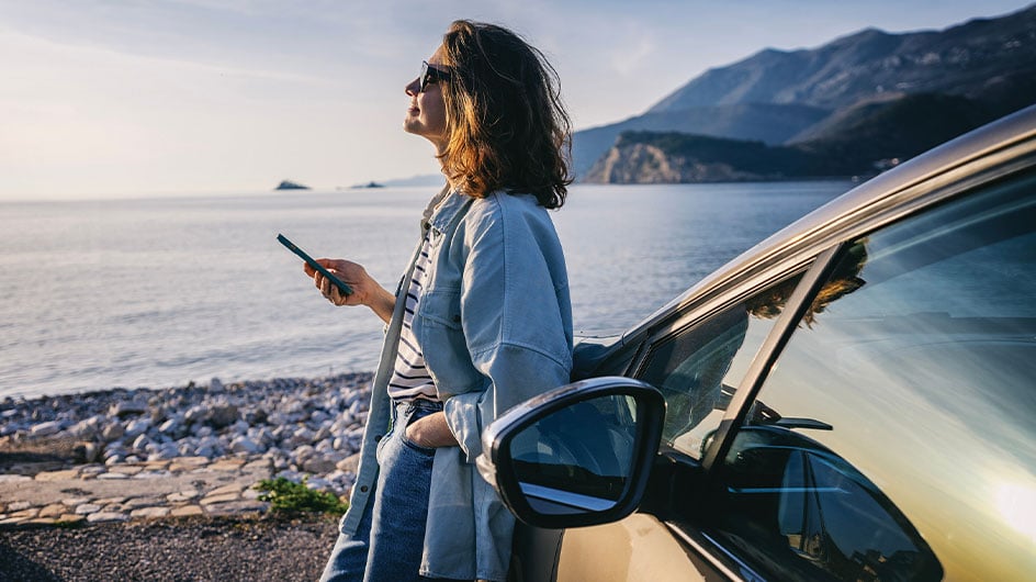 woman leaning against car using phone and admiring view