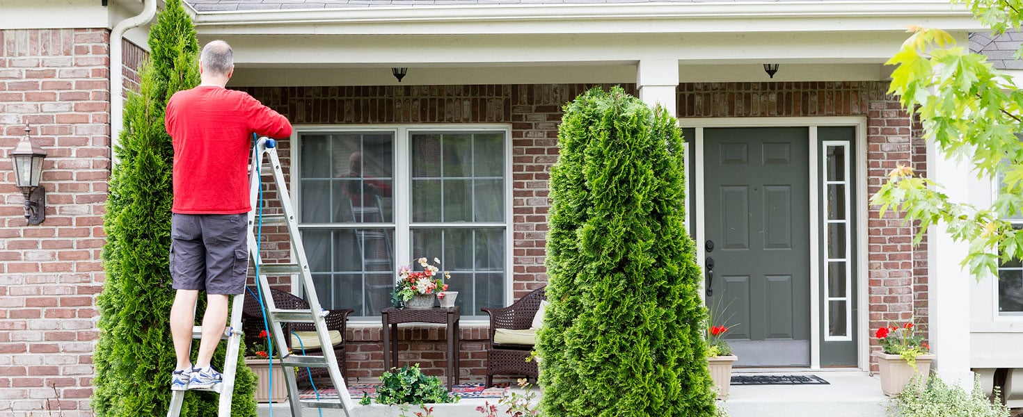 Man outside of his home, trimming a shrub, while on a ladder