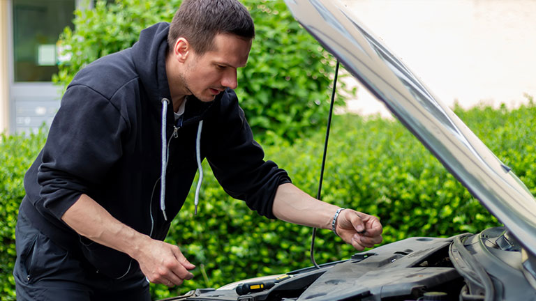The car broke down while traveling. A frustrated man near a broken car with an open hood far outside the city. A young guy looks under the hood of a car; by Jelena
