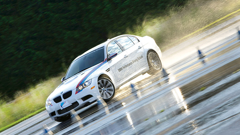 A white BMW car with "BMW Performance Driving School" signage skidding on a wet road during driver training, surrounded by cones.