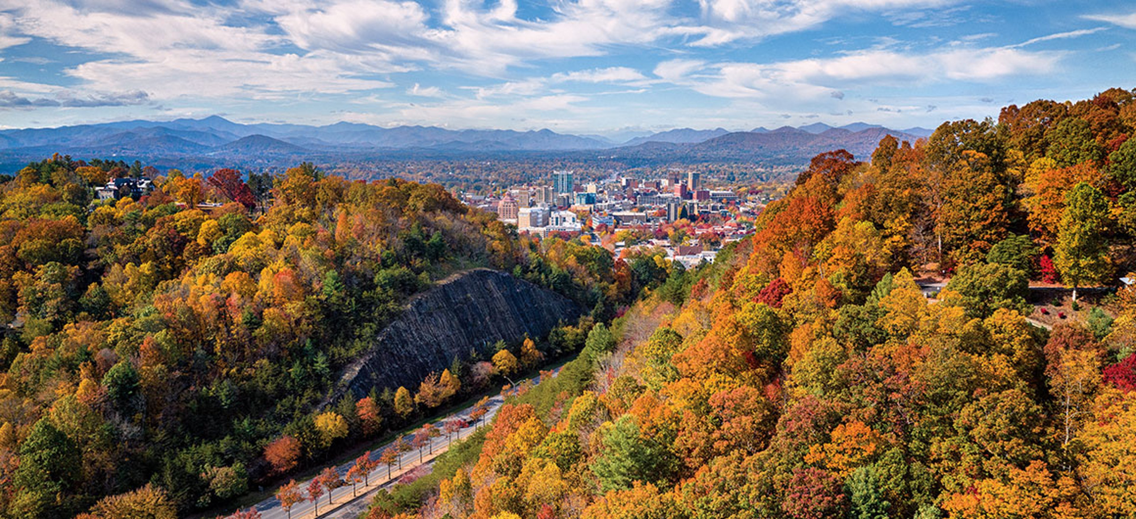 Aerial view of Asheville city surrounded by colorful autumn trees and distant mountains under a blue sky.