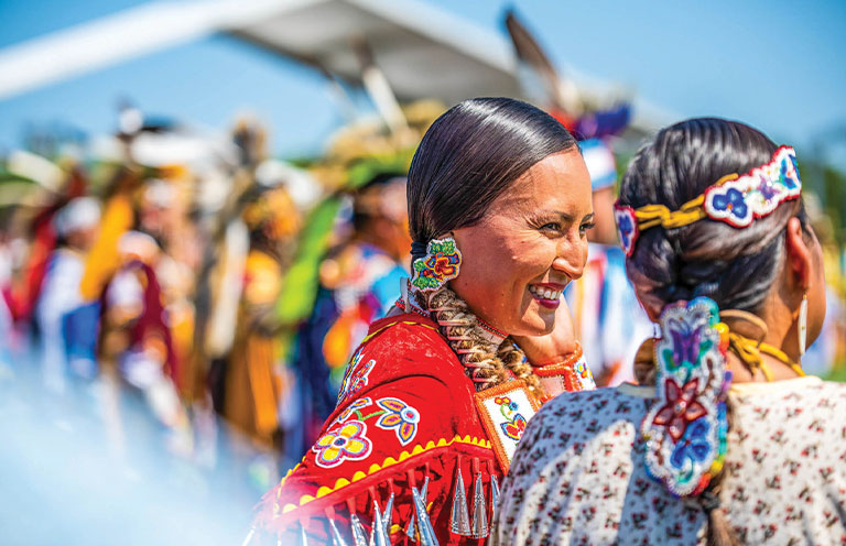 two women at a powwow in traditional attire smiling and having a conversation