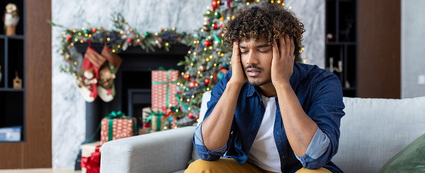 Severe hangover headache and migraines, man holding head sitting on sofa in living room for Christmas, African American man with curly hair near decorated Christmas tree.  By Liubomir