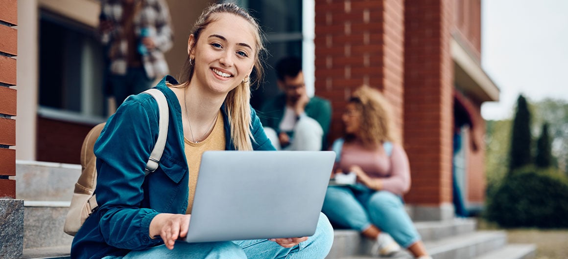 girl wearing bookbag sitting outside using laptop on college campus