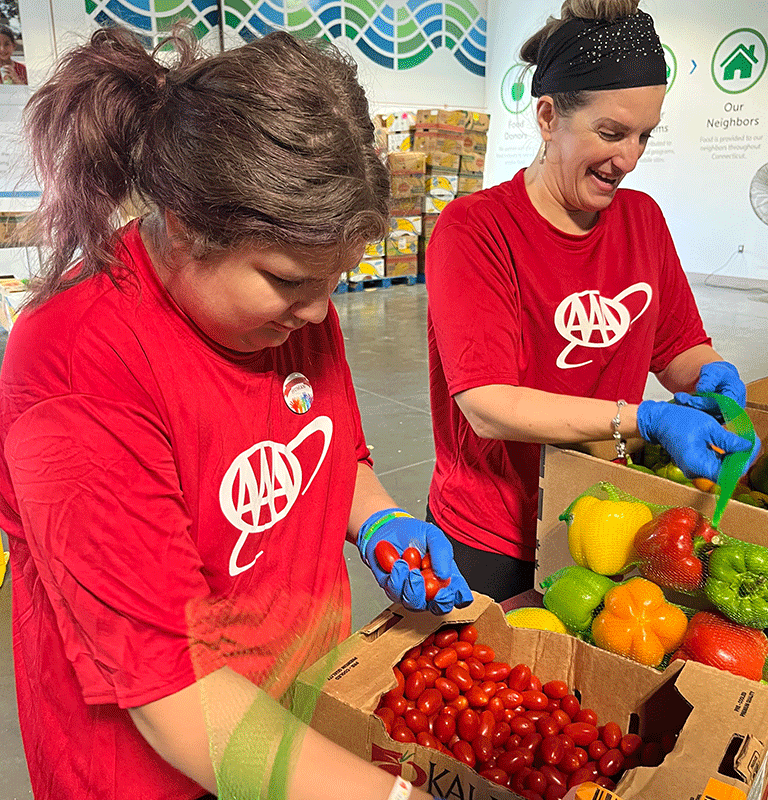 Two volunteers in red AAA shirts and blue gloves sort fresh produce at a food bank. One is placing cherry tomatoes into a mesh produce bag while the other smiles while handling a box of bell peppers. Colorful graphics and signs are visible on the wall behind them.