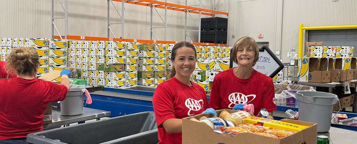 Two women in red AAA shirts stand at a packing station smiling in front of a box filled with canned goods. Behind them is a wall labeled “Nationwide Volunteer Center,” with rows of banana boxes stacked along the back wall and empty metal shelving overhead.