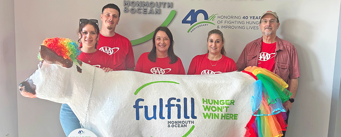 Five AAA team members in red shirts pose behind a large white statue of a cow decorated with a rainbow-colored tail and ears. The wall behind them displays the logo for Fulfill Monmouth & Ocean and the slogan “Hunger Won’t Win Here.”