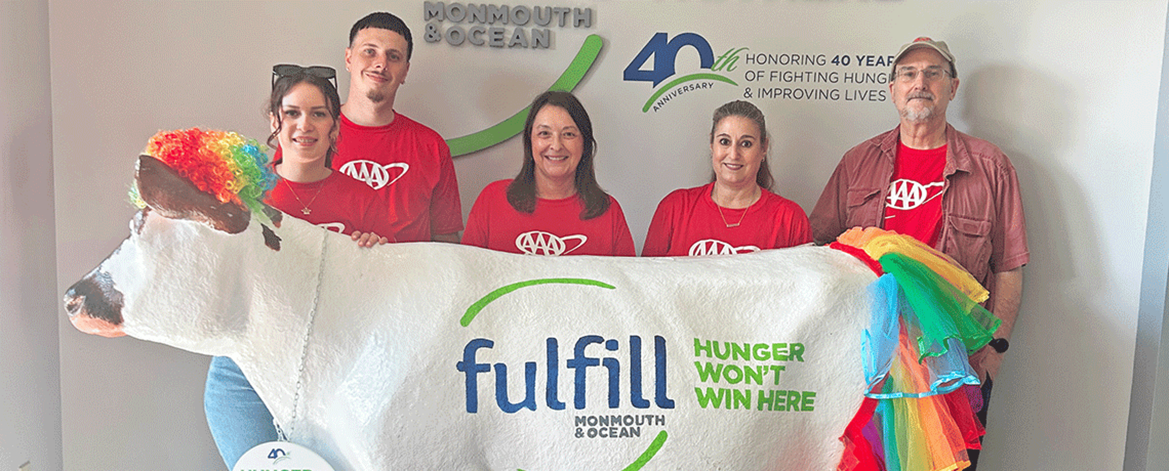 Five AAA team members in red shirts pose behind a large white statue of a cow decorated with a rainbow-colored tail and ears. The wall behind them displays the logo for Fulfill Monmouth & Ocean and the slogan “Hunger Won’t Win Here.”