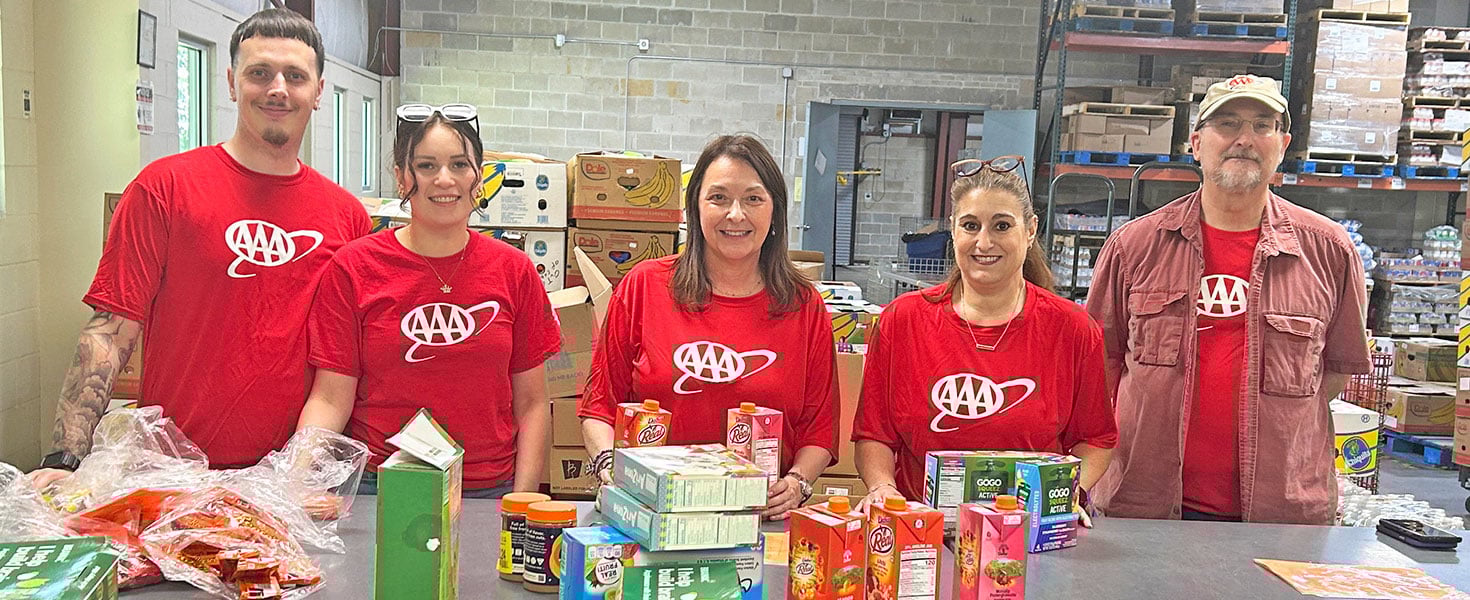 Five AAA team members in red shirts stand behind a table filled with assorted boxed snacks and fruit cups, including applesauce, fruit gels, juice boxes, and granola bars. They are smiling and posing in a warehouse setting with shelves and banana boxes in the background.