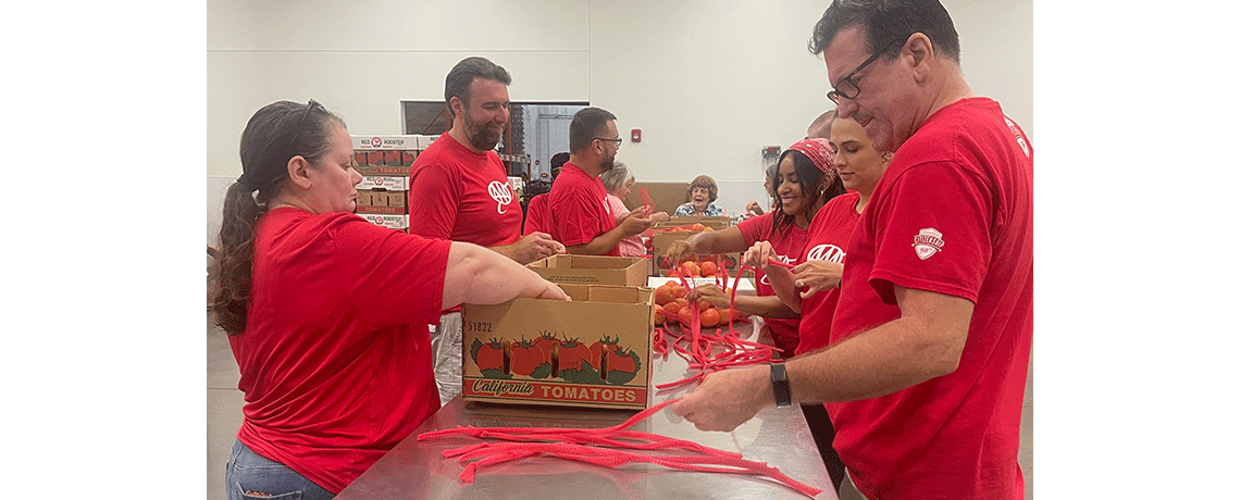 A group of volunteers in red AAA shirts gather around a table organizing red produce and plastic ties. Boxes labeled “California Tomatoes” are on the table, and volunteers are working together in a large, well-lit warehouse.