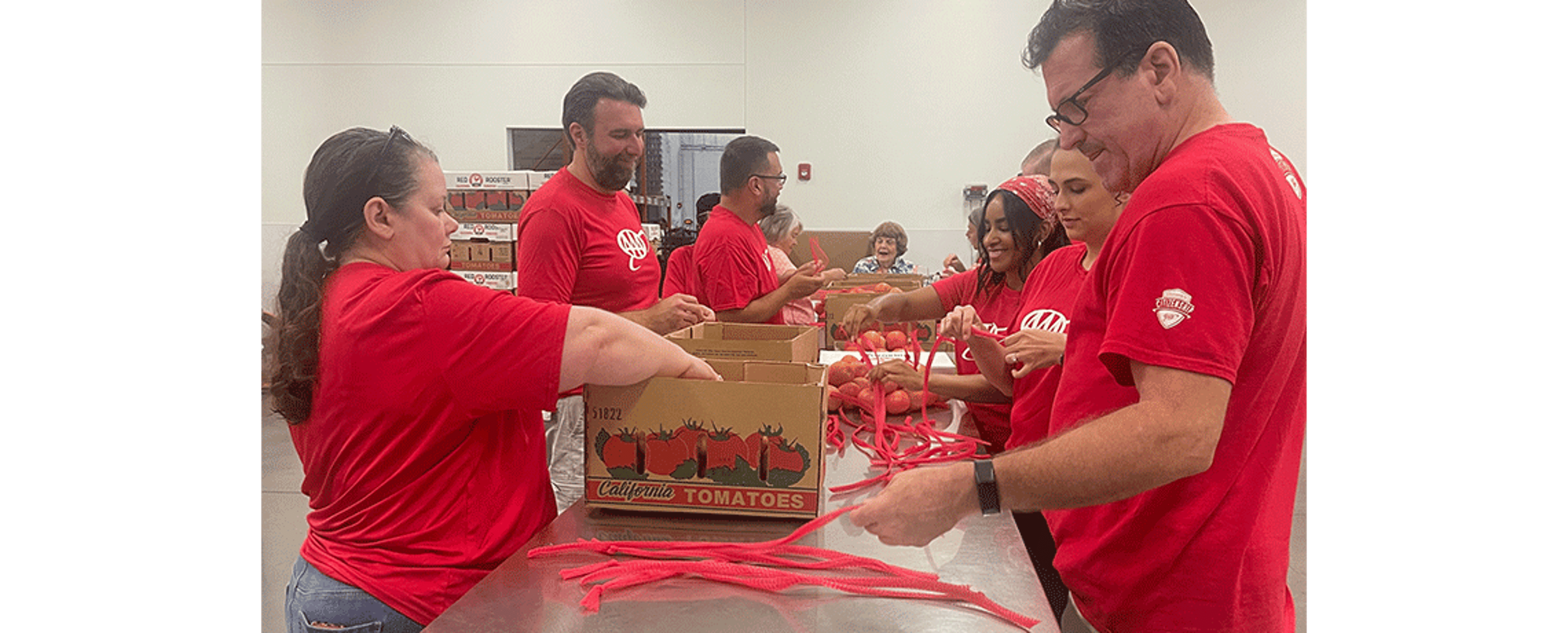 A group of volunteers in red AAA shirts gather around a table organizing red produce and plastic ties. Boxes labeled “California Tomatoes” are on the table, and volunteers are working together in a large, well-lit warehouse.