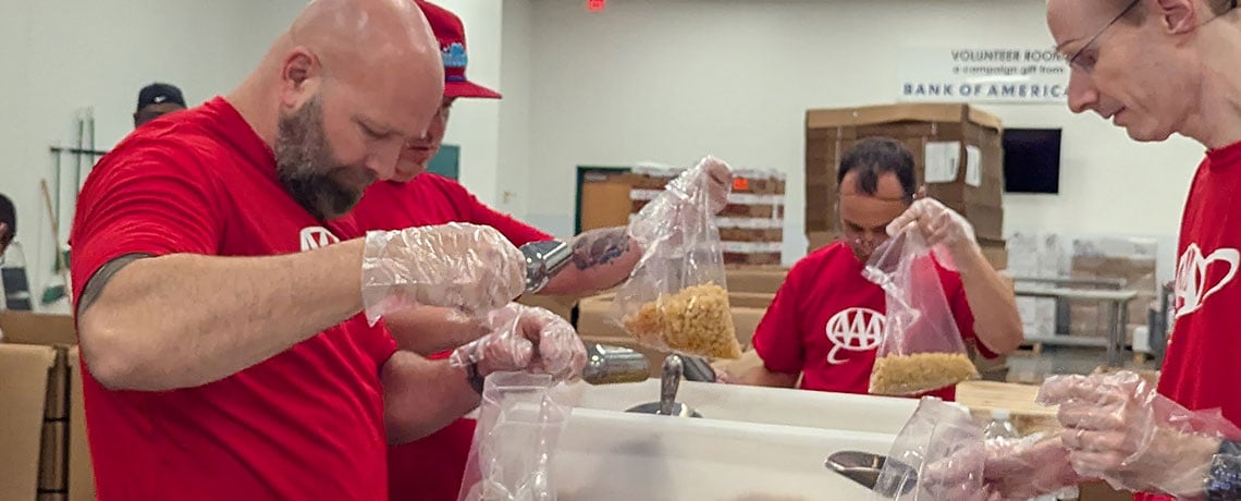 Four volunteers in red AAA shirts are wearing gloves and scooping dry cereal or pasta into clear plastic bags using metal scoops. The workspace includes metal tables and food scales.