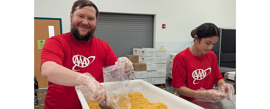 A bearded man in a red AAA shirt smiles while scooping dry pasta into a plastic bag at a volunteer station. A young woman next to him works on sealing bags. Boxes labeled “snacks” are stacked in the background